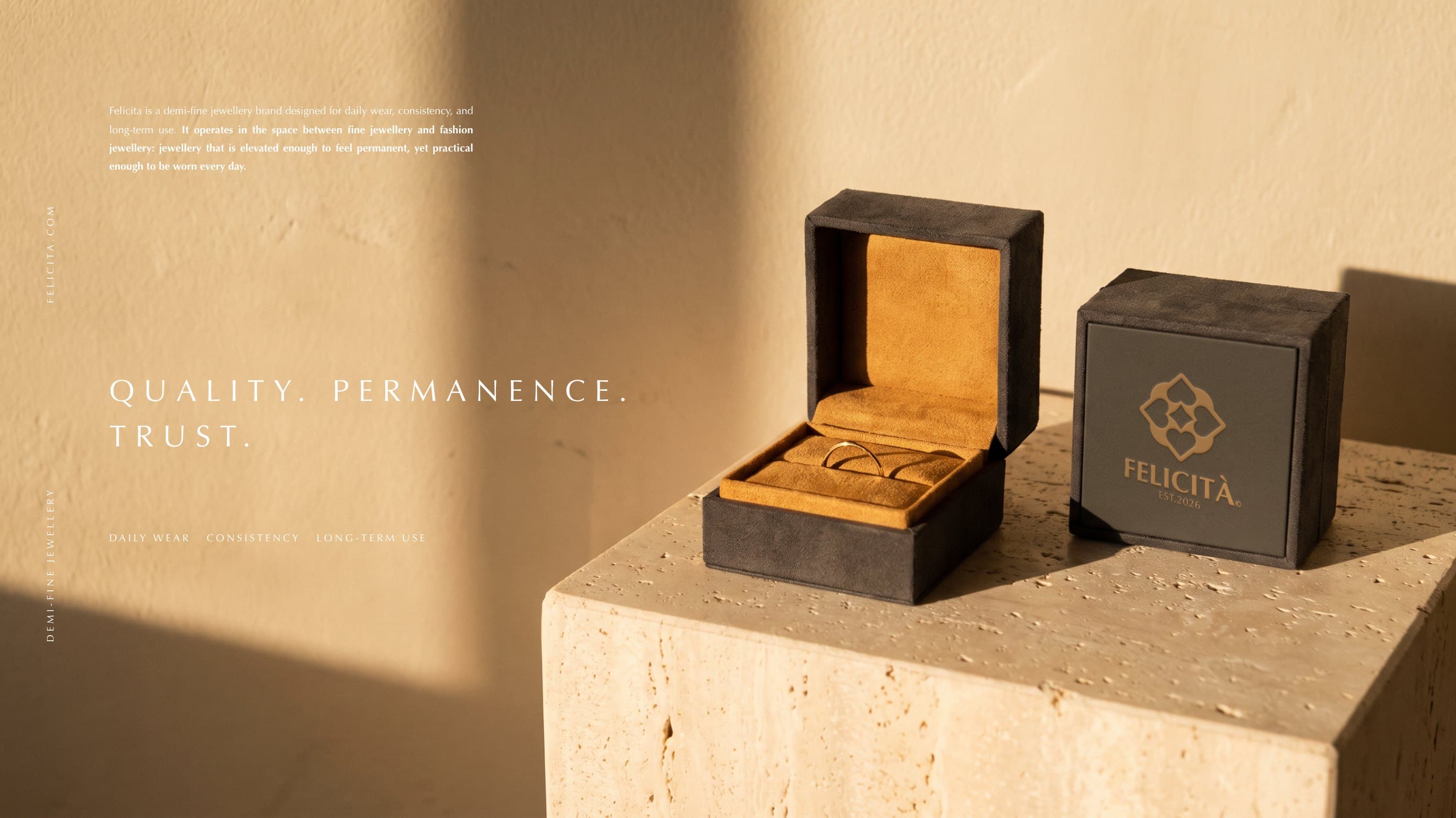 Two charcoal Felicità ring boxes on travertine in late sun, the emblem and wordmark foiled in warm gold: Quality. Permanence. Trust.