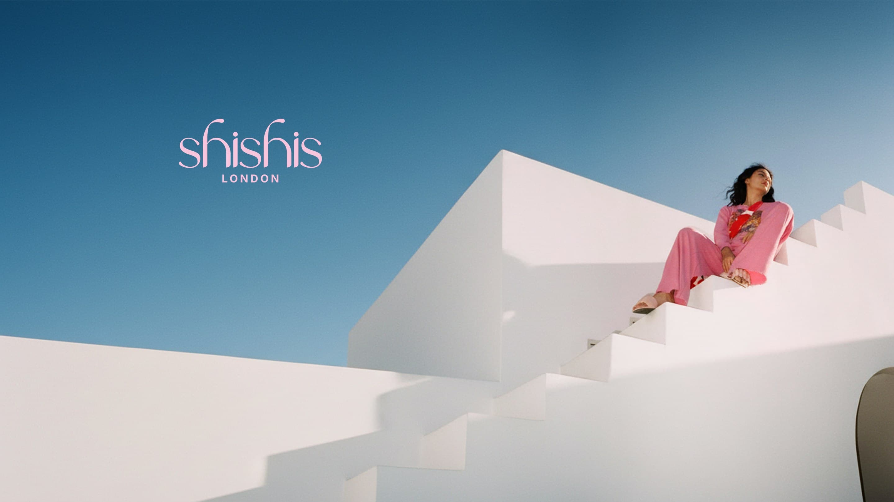 A woman in pink sits high on white stairs against a blue sky, the shishis London wordmark in the sky